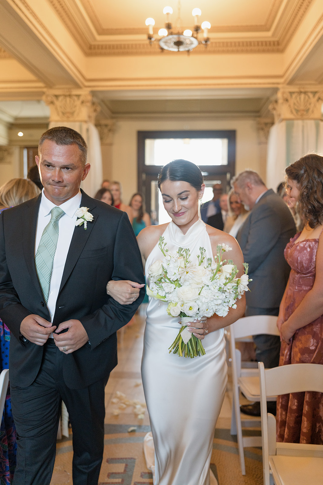 Bride walking down the aisle with her father beneath the chandelier at her Oklahoma Hall of Fame wedding ceremony in Oklahoma City