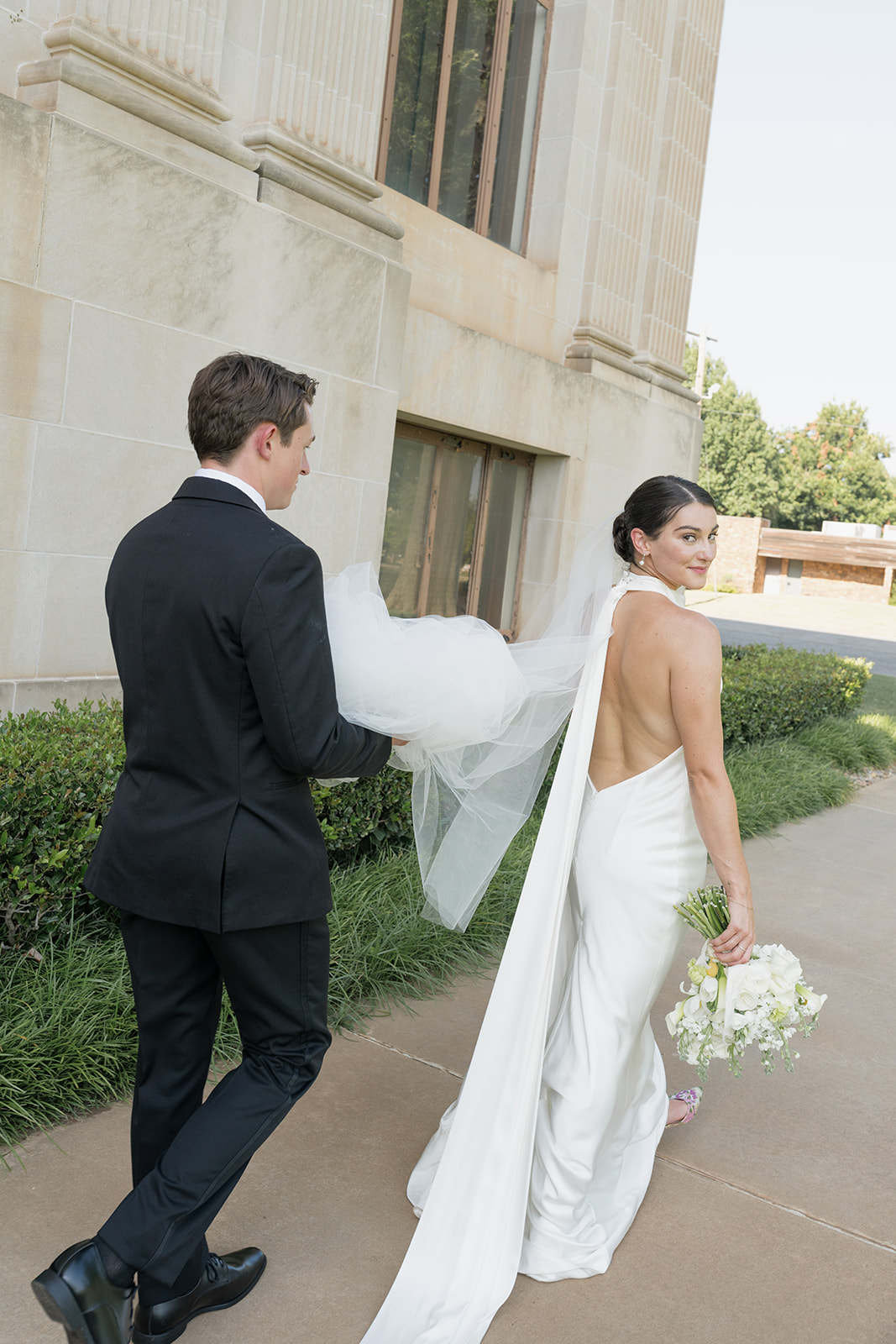Playful portrait of the groom carrying the bride’s veil as they walk around the Oklahoma Hall of Fame grounds in Oklahoma City