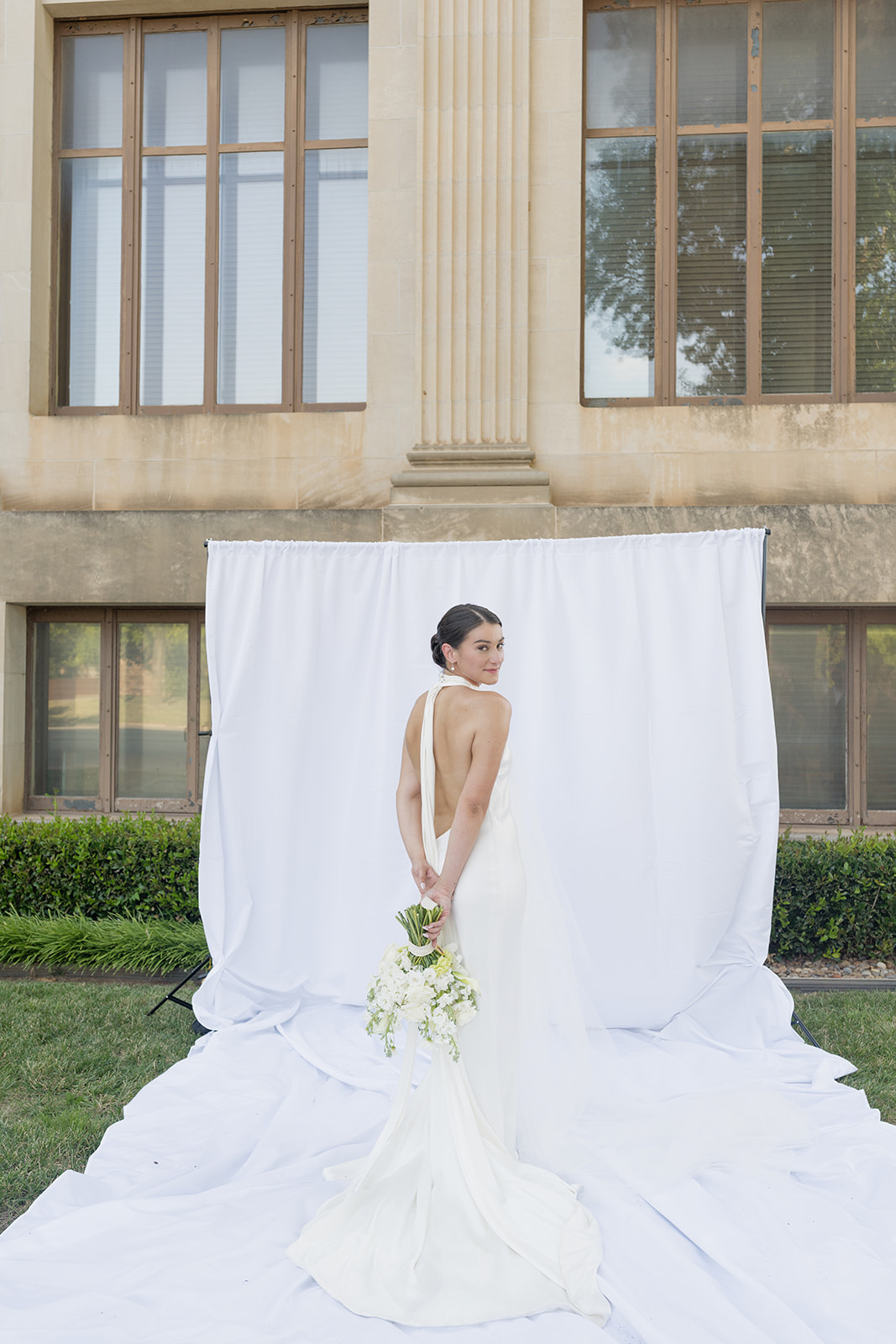 Bridal portrait showing the open back of her satin gown with a white backdrop at the Oklahoma Hall of Fame in Oklahoma City